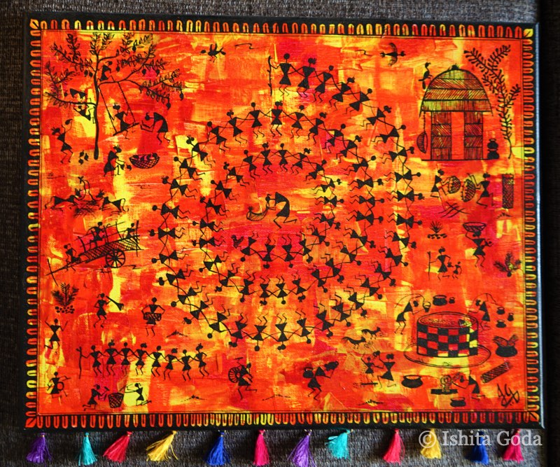 Warli-style painting: black stick figures dancing in a large spiral among huts, trees and animals on a fiery orange and red background, with coloured tassels along the bottom.